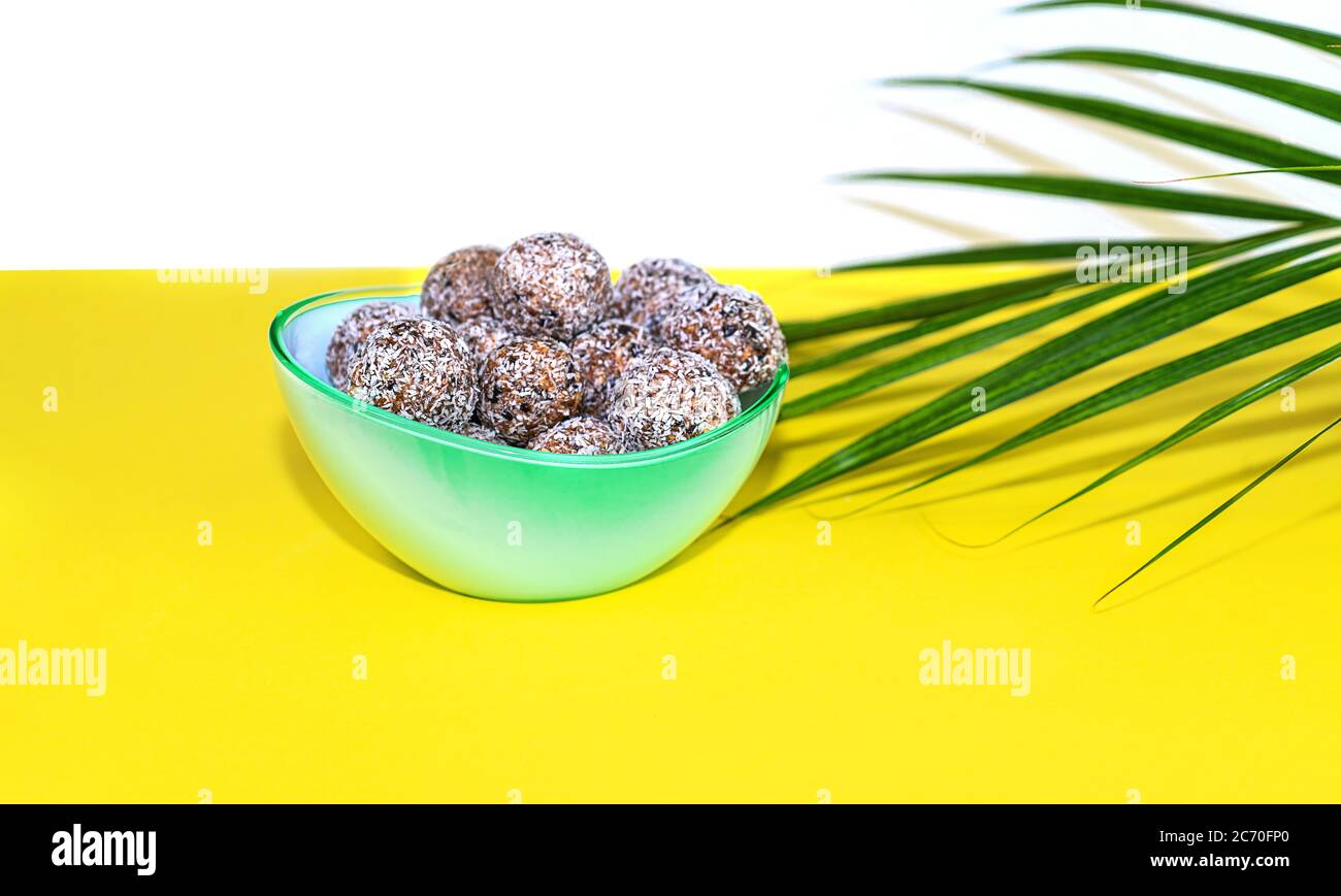 Protein Erdnussbutter Energie Bisse oder Energiebälle. Hausgemachte rohe Schokoladentrüffel mit Nüssen und in Kokosflocken auf Teller mit Palmblatt auf gelbem Hintergrund. Stockfoto