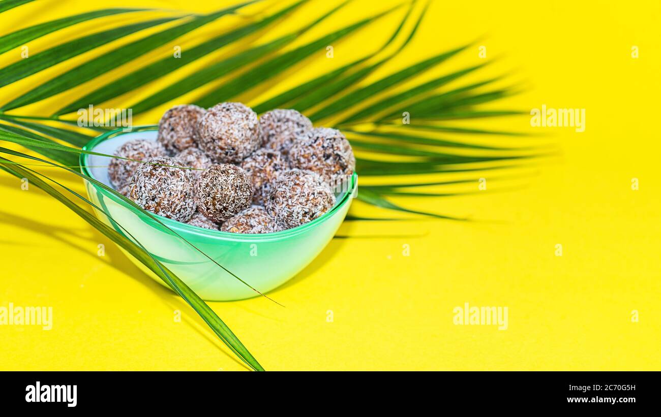 Protein Erdnussbutter Energie Bisse oder Energiebälle. Hausgemachte rohe Schokoladentrüffel mit Nüssen und in Kokosflocken auf Teller mit Palmblatt auf gelbem Hintergrund. Stockfoto