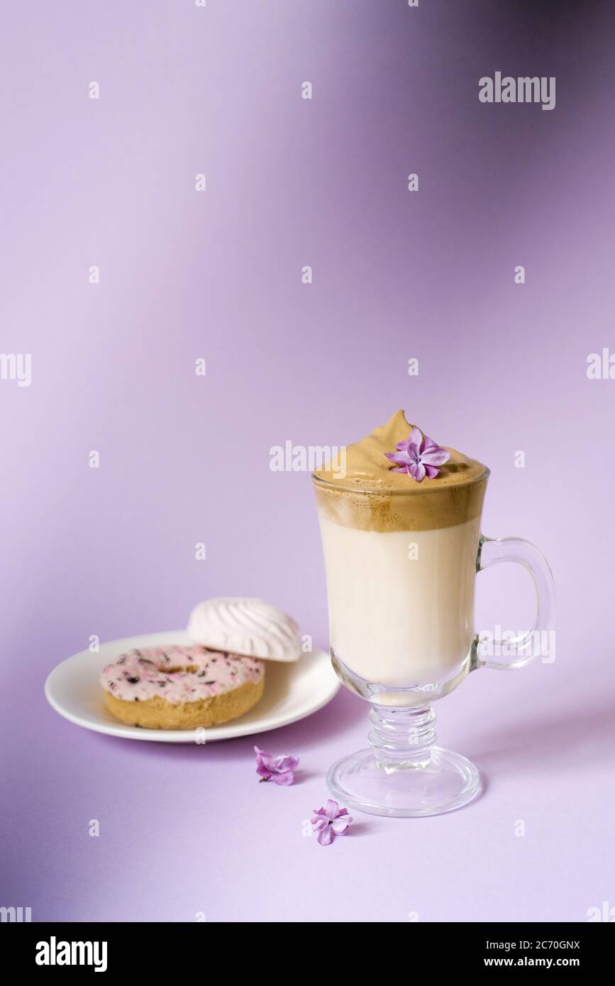 Schöne dalgona trinken einen schaumigen Kaffee in einem transparenten Becher und Blumen aus Flieder auf einem violetten Hintergrund. Süßigkeiten auf einem Teller: Donat und zarter Marschall Stockfoto