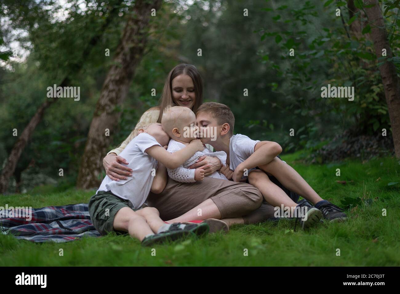 Junge Mutter mit drei kleinen Kindern, die im Sommer Picknick machen. Große Cousine umarmen und küssen ihre jüngere Schwester Stockfoto