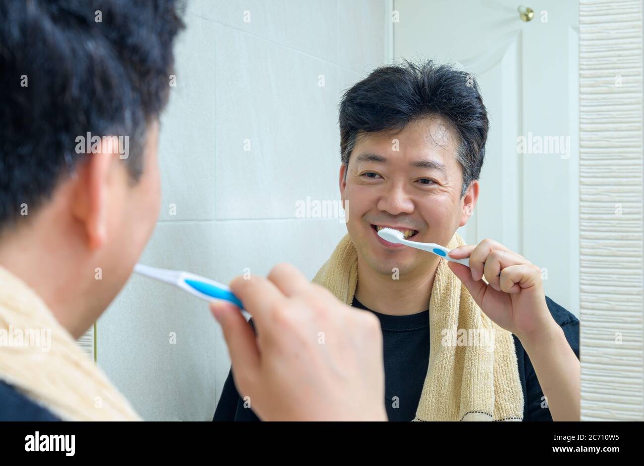 Ein Mann mittleren Alters, der ein Handtuch um den Hals trägt und im Badezimmer Zähne putzt. Stockfoto