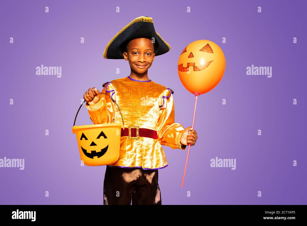 Kleiner netter schwarzer Junge im Halloween Kostüm zeigt lächelnd und stehend über purpurem Hintergrund und hält gruseligen Bonbons Eimer Stockfoto