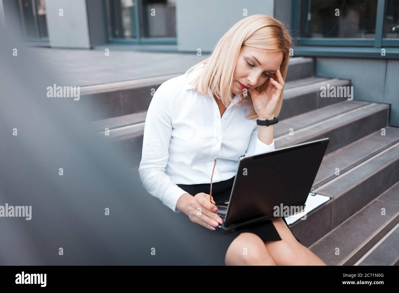 Müde Büroangestellte sitzt auf der Treppe hinter einem Laptop und hält ihren Kopf. Überarbeitungskonzept Stockfoto