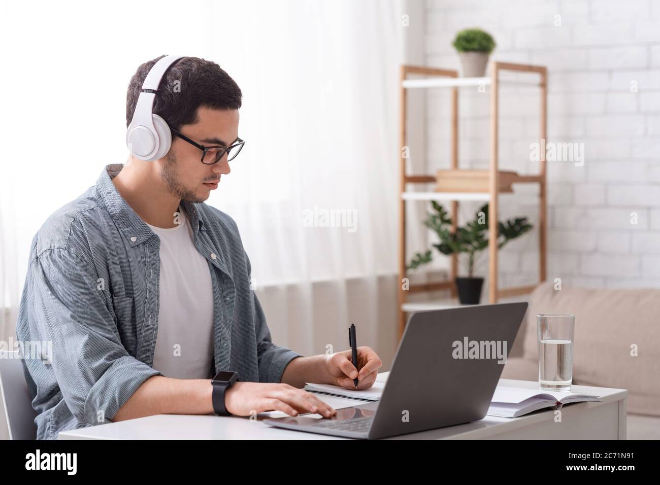 Moderne Gadgets für Arbeit und Studium online. Kerl mit großen Kopfhörern funktioniert auf Laptop und macht Notizen in Notebook Stockfoto Moderne Gadgets für Arbeit und Studium online. Kerl mit großen Kopfhörern funktioniert auf Laptop und macht Notizen in Notebook Stockfoto