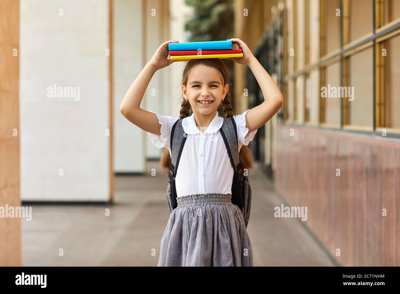 Zurück zur Schule. Schulmädchen in Uniform hält Haufen von Büchern auf ihrem Kopf in der Nähe des Schulgebäudes. Stockfoto