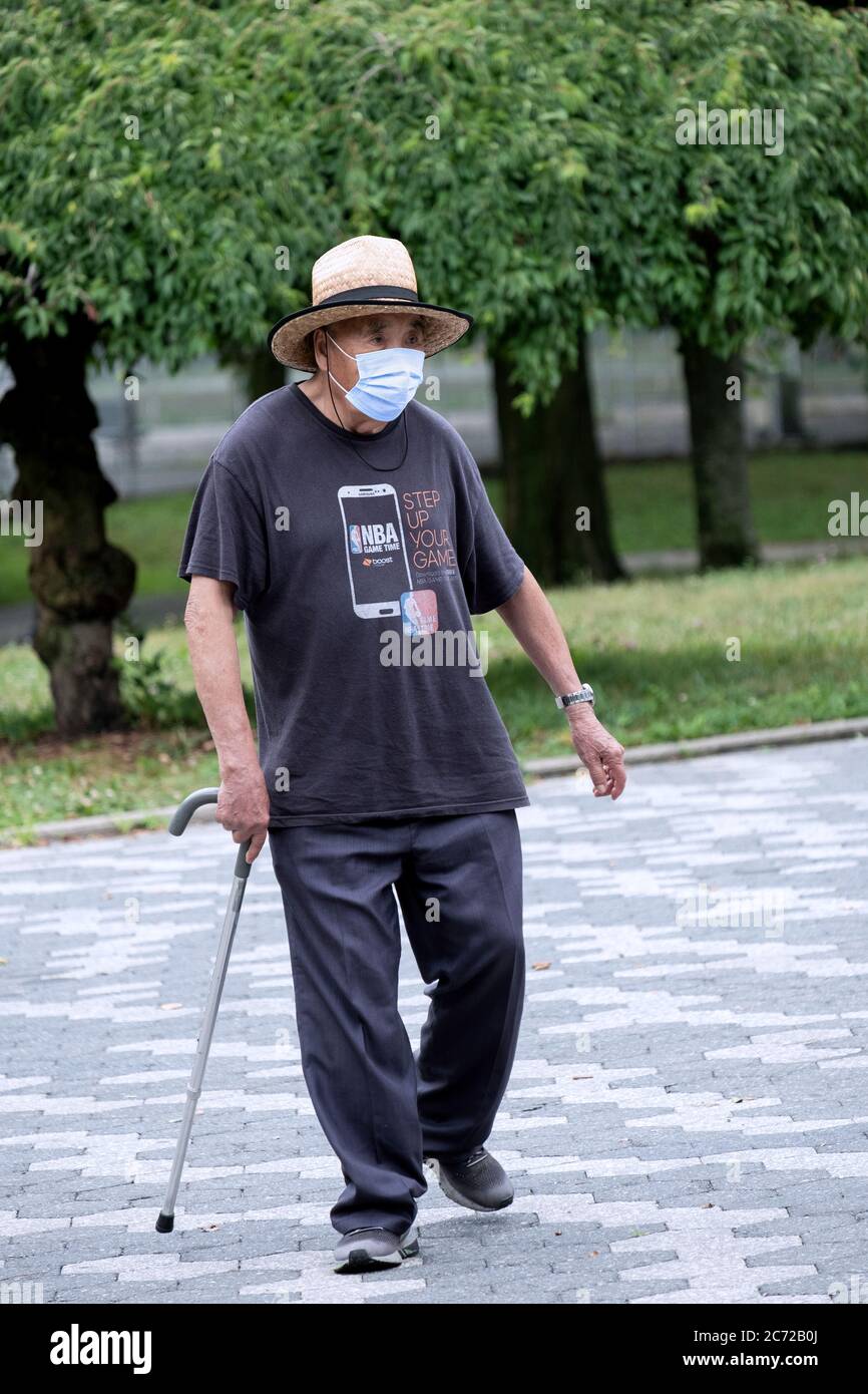 Ein asiatischer amerikanischer Senioren führt mit einem Stock und unter dem Tragen einer chirurgischen Maske Spaziergänge durch. In Kissena Park, Flushing, Queens, New York City. Stockfoto