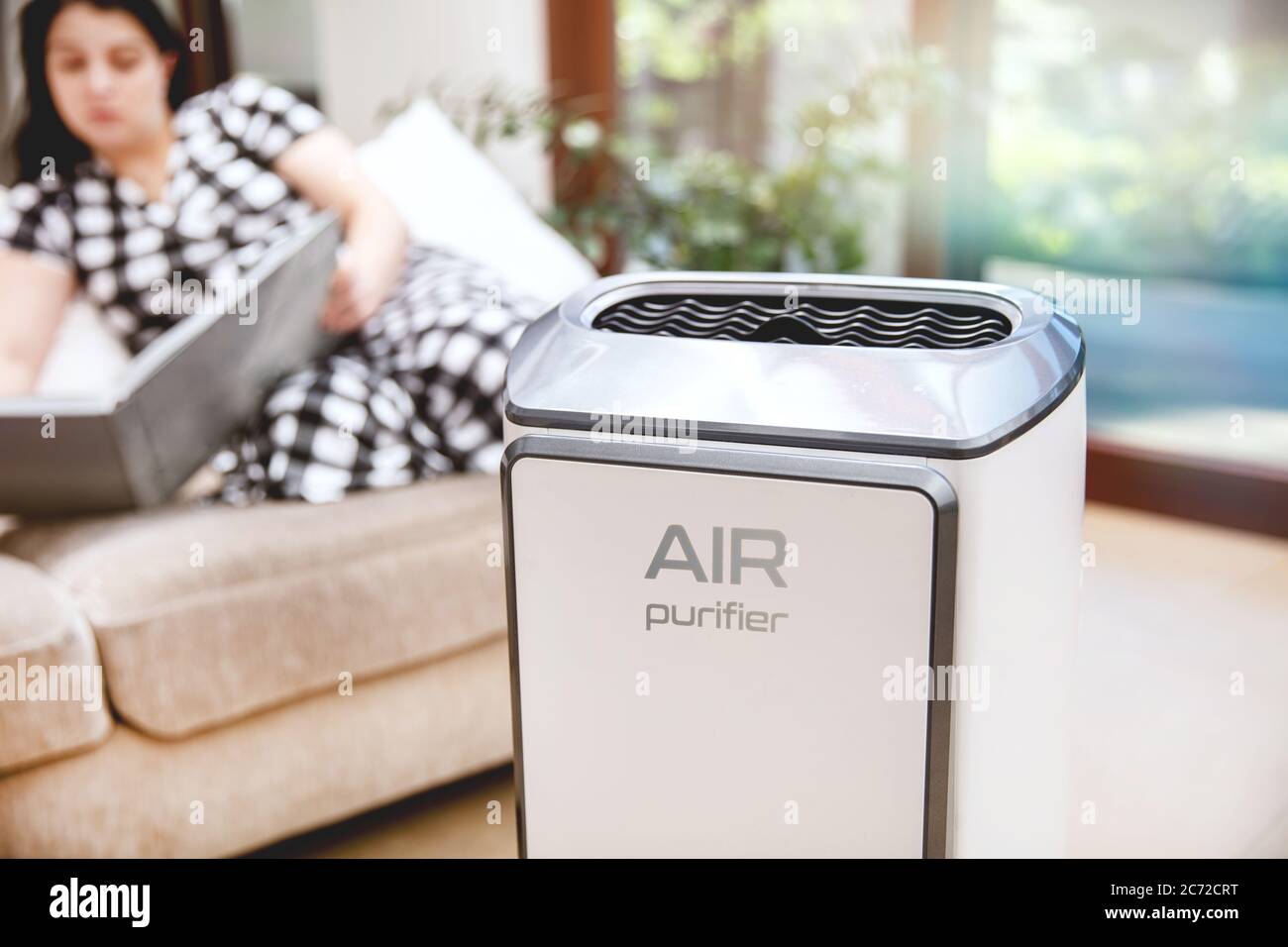 Luftreiniger reinigt die Luft. Moderner Luftreiniger reinigt die Luft im Wohnzimmer mit Frau, die ein Buch auf der Couch im Hintergrund liest. Logo auf dem Stockfoto