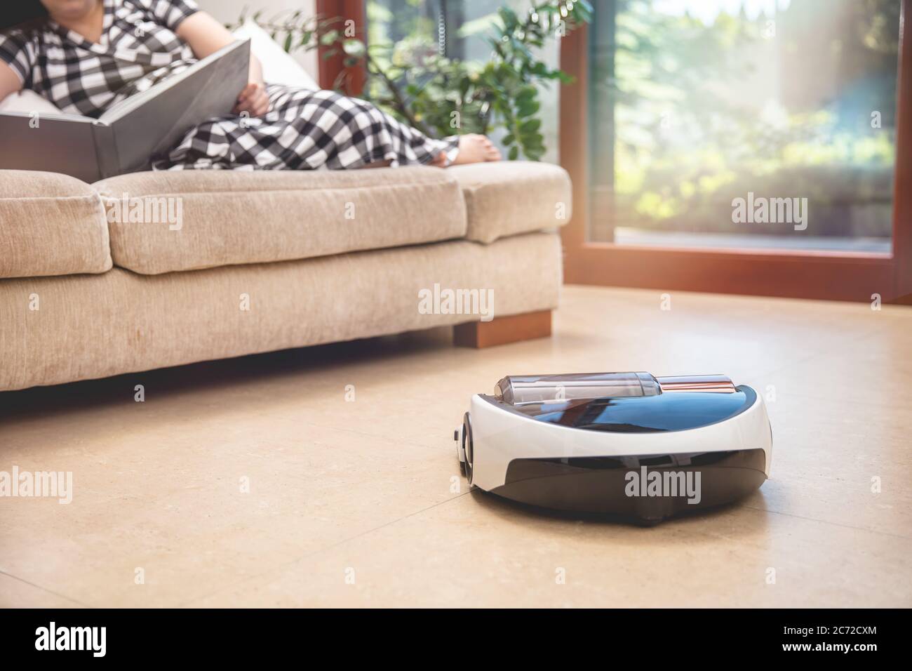 Roboter-Staubsauger, der in modernen Wohnheims arbeitet. Round hoover reinigt das Wohnzimmer selbst, während Frau auf der Couch liegt und sich entspannen Stockfoto