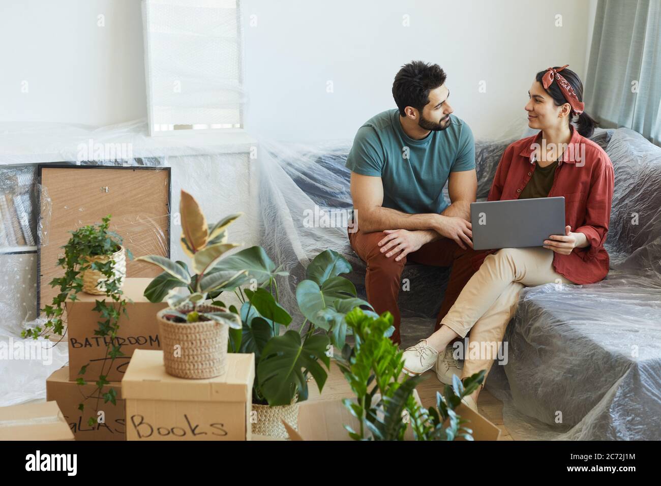Junges Paar, das sich unterhielt und mit einem Laptop auf dem Sofa in ihrer neuen Wohnung saß Stockfoto