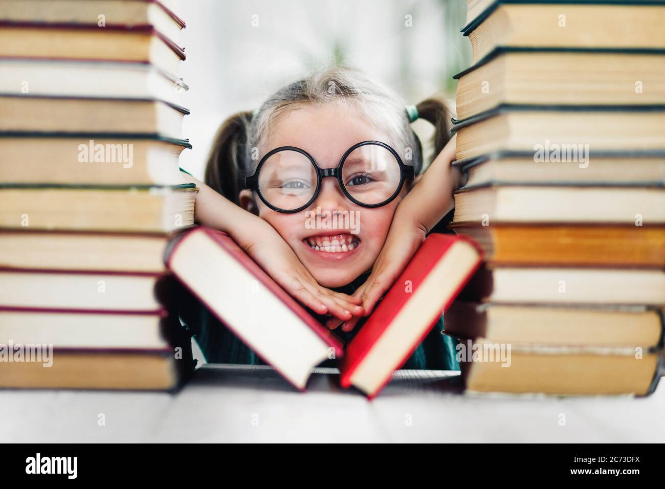 Mädchen im Vorschulalter mit Grübchen in großen lustigen Gläsern zwischen einem Stapel von Büchern Stockfoto