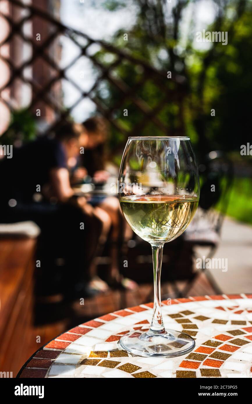 Ein Glas Weißwein auf einem Mosaiksteintisch einer Café-Terrasse an einem sonnigen Tag, Menschen im Hintergrund Stockfoto