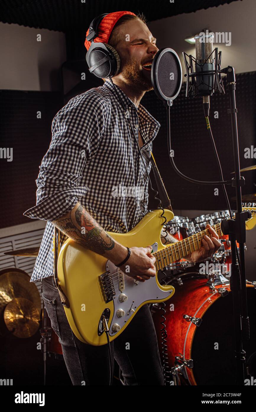 Junge kaukasische Gitarrist gleichzeitig Gitarre spielen und singen ein Lied im Aufnahmestudio. Schöner Kerl in Red hat emotional singen Song, scharf auf mu Stockfoto