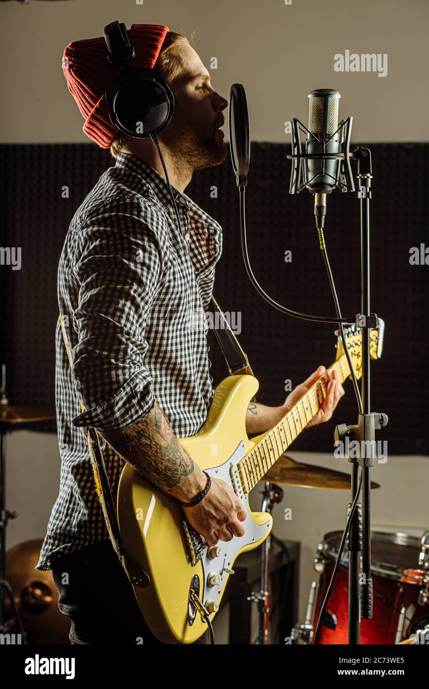 Junge kaukasische Gitarrist gleichzeitig Gitarre spielen und singen ein Lied im Aufnahmestudio. Schöner Kerl in Red hat emotional singen Song, scharf auf mu Stockfoto