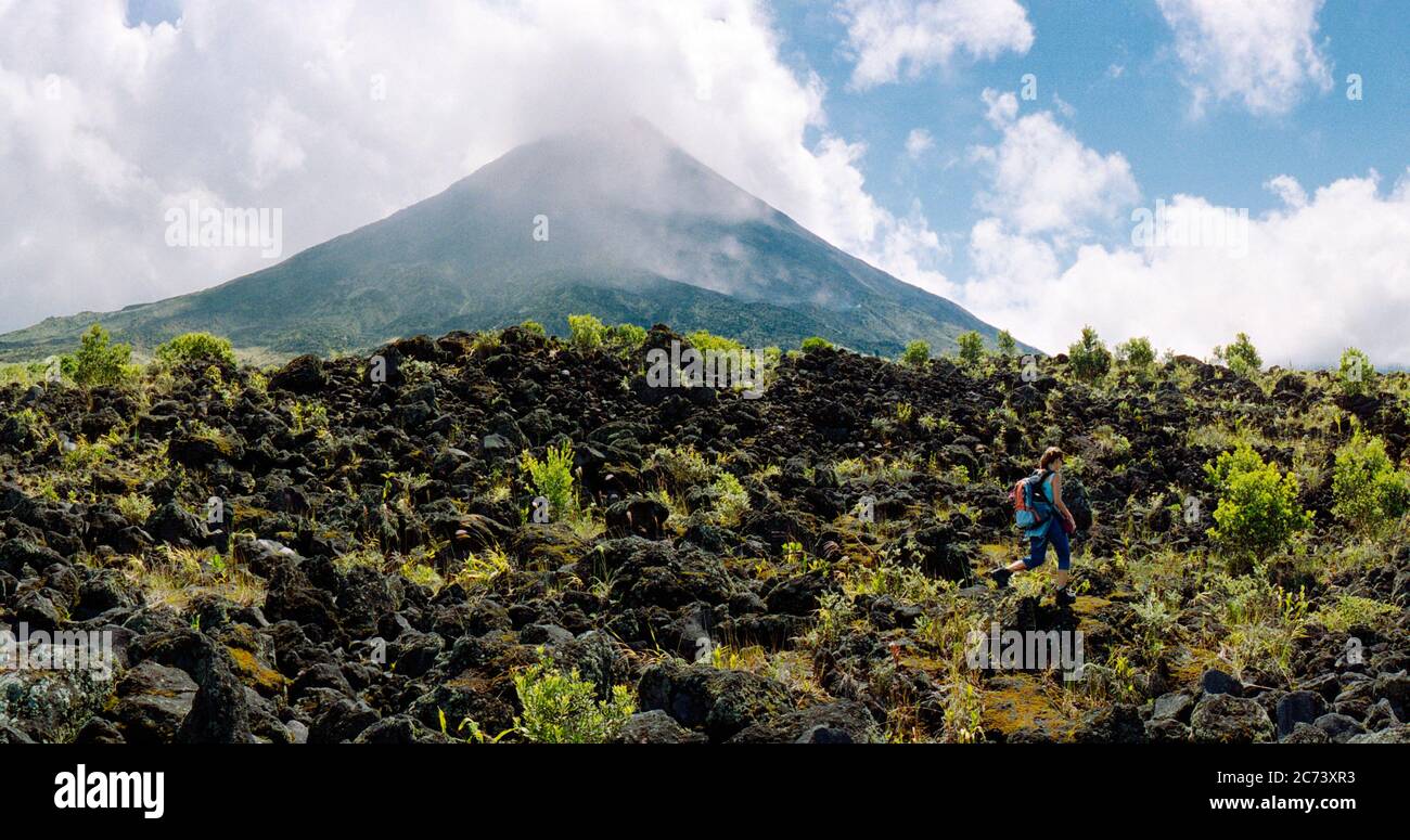 Weite Panoramaaussicht auf den Vulkan arenal in einem Nationalpark in Costa rica mit einigen Wolken. Eine Frau läuft im Vordergrund auf Lavagesteinen mit Stockfoto Weite Panoramaaussicht auf den Vulkan arenal in einem Nationalpark in Costa rica mit einigen Wolken. Eine Frau läuft im Vordergrund auf Lavagesteinen mit Stockfoto