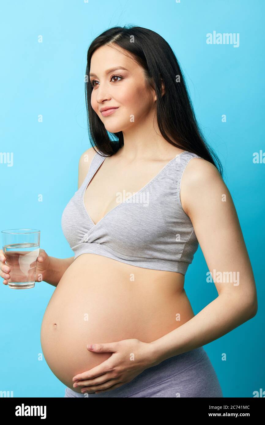 Junge schwangere kaukasische Modell mit langen schwarzen Haaren hält ein Glas Wasser. Unterstützung des Wasserhaushaltssangleichs, so notwendig für sie und ihr erwartetes Baby. Stu Stockfoto