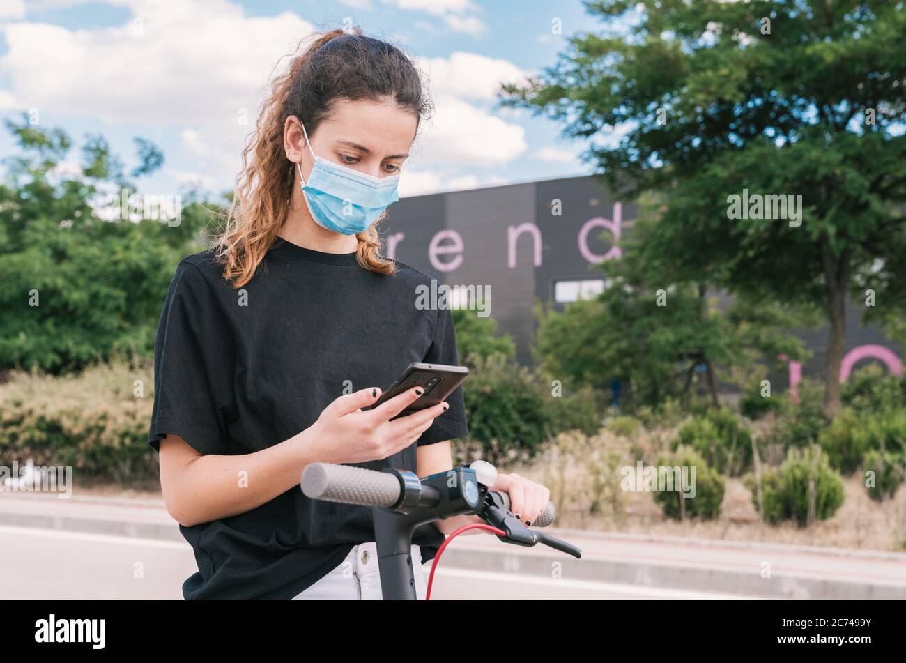 Neue normale. Mädchen mit einer Maske mit ihrer Handy-App, um einen Verleih Elektroroller zu aktivieren. Teenager mit einer Maske mit einem Elektroroller. Junger Bursche Stockfoto