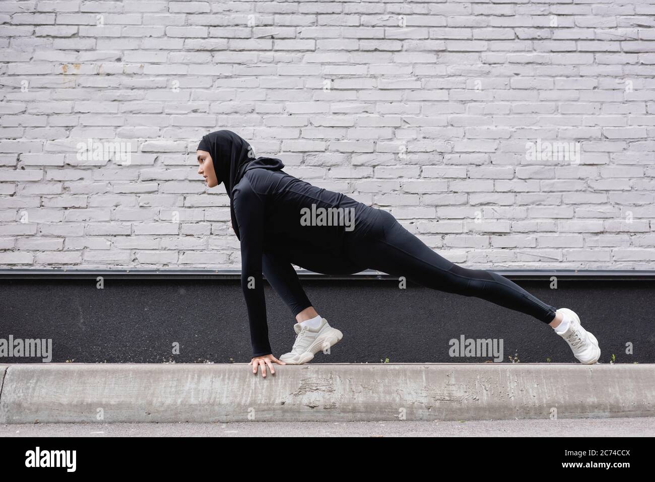 Seitenansicht der arabischen Sportlerin in Hijab, die sich in der Nähe einer Ziegelmauer erstreckt Stockfoto