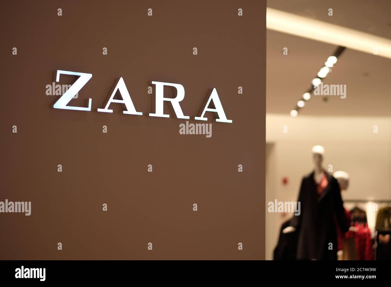 Nahaufnahme des beleuchteten ZARA-Logos an der Wand. Verwischen Sie Kleidung als Hintergrund. Eine spanische Modemarke. Stockfoto