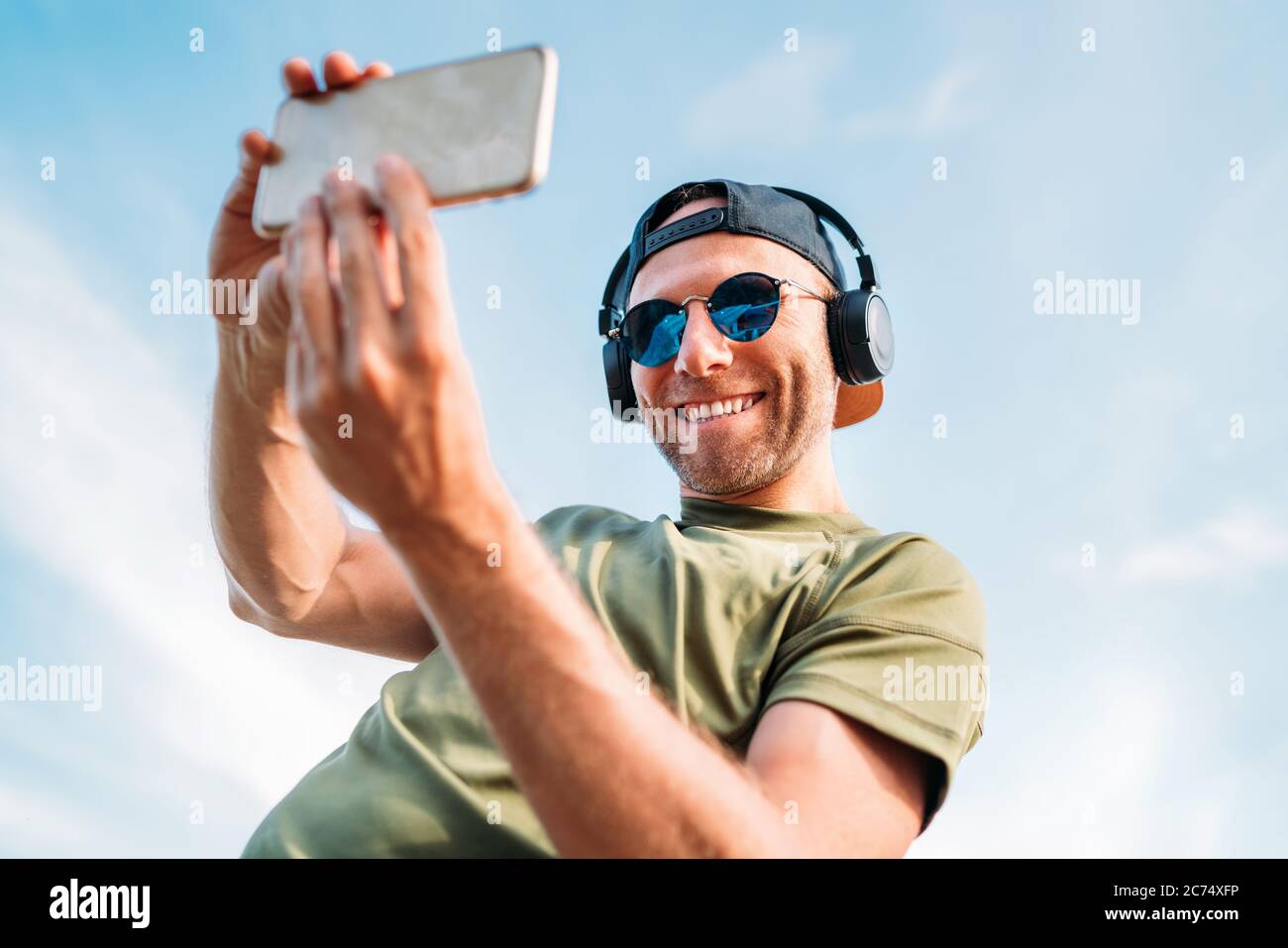 Bärtiger Mann mit Baseballmütze, kabellosen Kopfhörern und blauer Sonnenbrille lächelt fröhlich und macht mit einem modernen Smartphone ein Selfie-Bild. Stockfoto