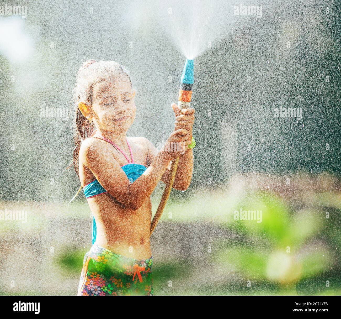 Nettes kleines Mädchen streut ein Wasser für sich aus dem Schlauch, macht einen Regen. Vergnügen für heiße Sommertage Stockfoto