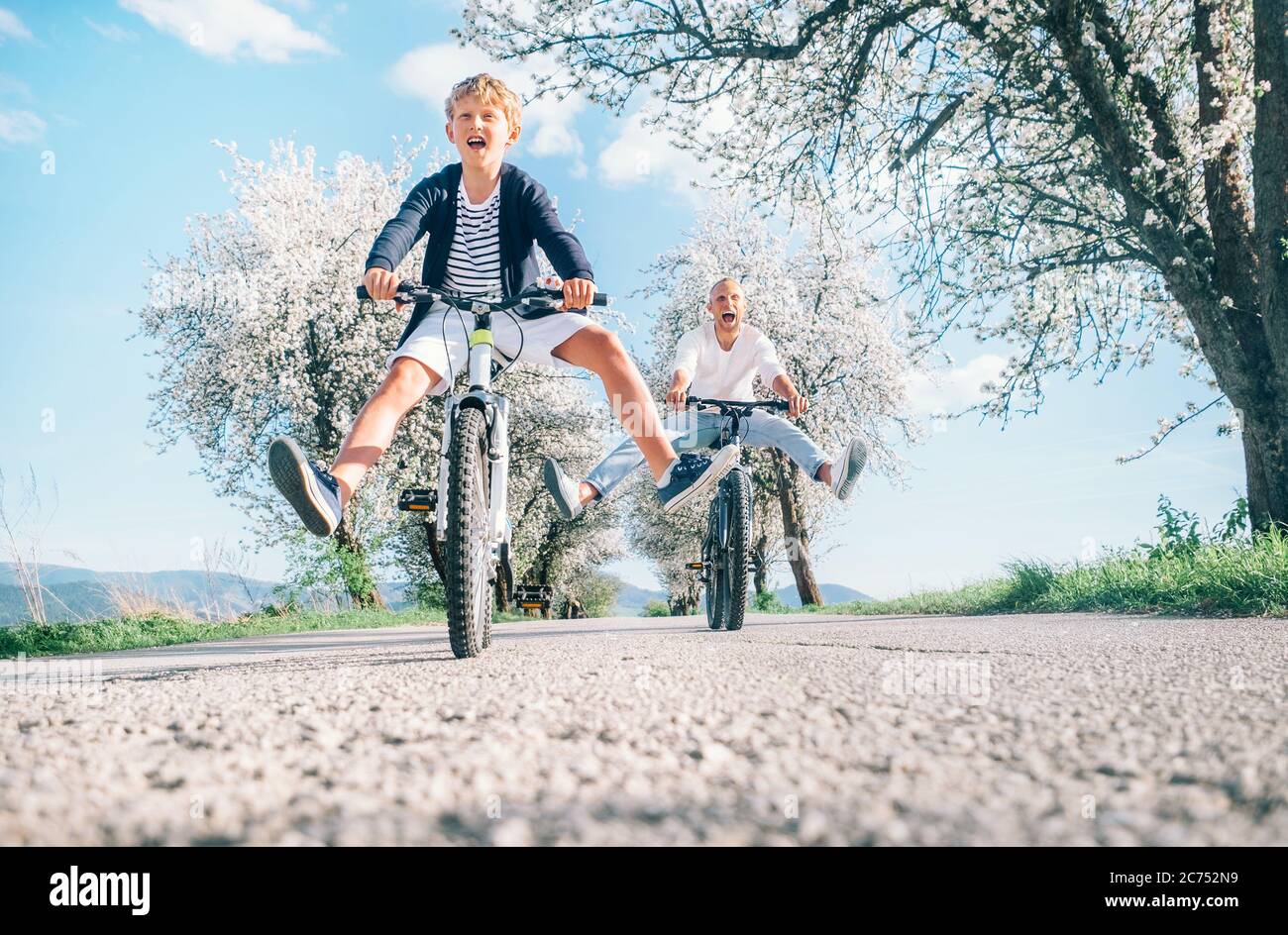 Vater und Sohn haben Spaß, breiten Beinen und Schreien beim Radfahren auf der Landstraße unter Blütenbäumen. Gesundes sportliches Lifestyle-Konzept Stockfoto