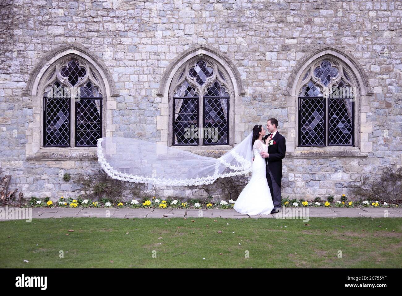 Brautpaar mit weißem Brautkleid und Schleier, der vor gotischen Fenstern im Wind weht oder weht Stockfoto