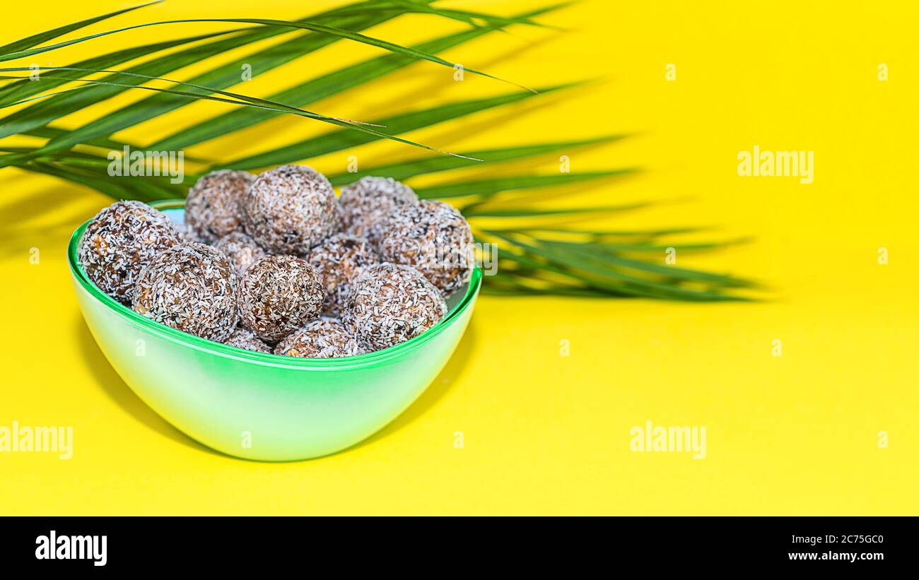 Protein Erdnussbutter Energie Bisse oder Energiebälle. Hausgemachte rohe Schokoladentrüffel mit Nüssen und in Kokosflocken auf Teller mit Palmblatt auf gelbem Hintergrund. Stockfoto