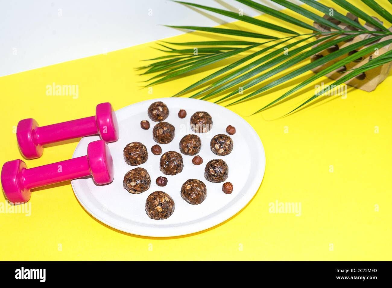 Protein Erdnussbutter Energie Bisse oder Energiebälle. Hausgemachte rohe Schokoladentrüffel mit Nüssen auf Teller mit Hanteln und Palmblatt auf gelbem Hintergrund. Stockfoto