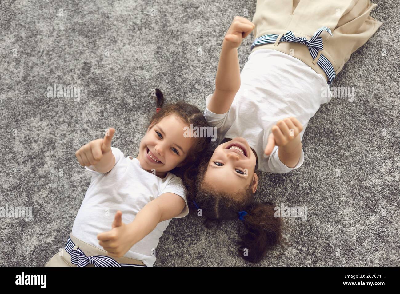 Zwillingsschwestern Kind zeigt Daumen nach oben Geste, während auf weichem Teppich liegen, Draufsicht. Fröhliche Geschwister verbringen Zeit zusammen Stockfoto