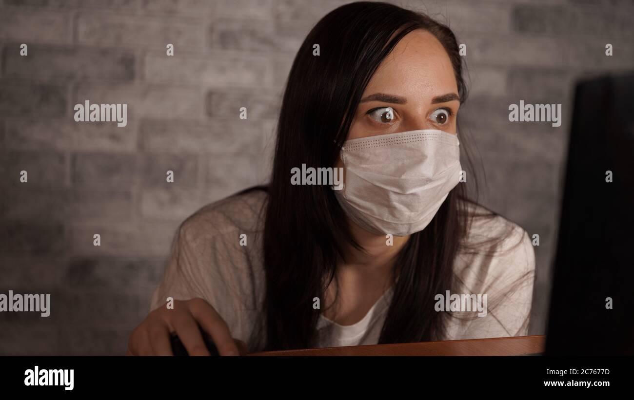 Eine Frau in einer medizinischen Maske erfährt mit einem Laptop aus dem Internet Neuigkeiten über das Coronavirus. Frau ist besorgt über seine Gesundheit. Die Epidemie Von Stockfoto