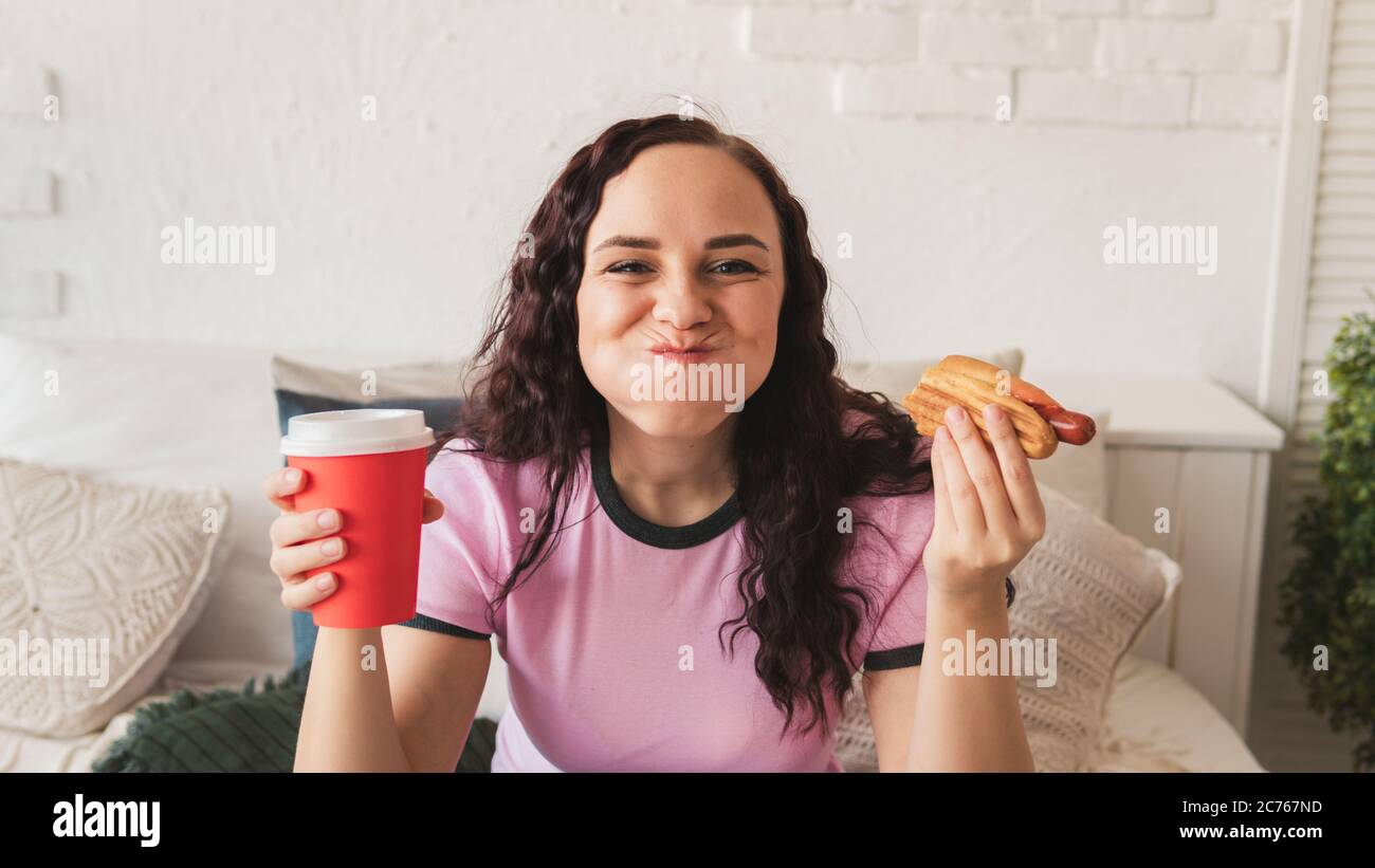 Eine junge Frau im Bett, die einen Hot Dog isst und Kaffee trinkt. Weibchen isst einen saftigen Hot Dog in einem gemütlichen Schlafzimmer. Konzept: Versuchung auf Diät und Fast-Food Stockfoto