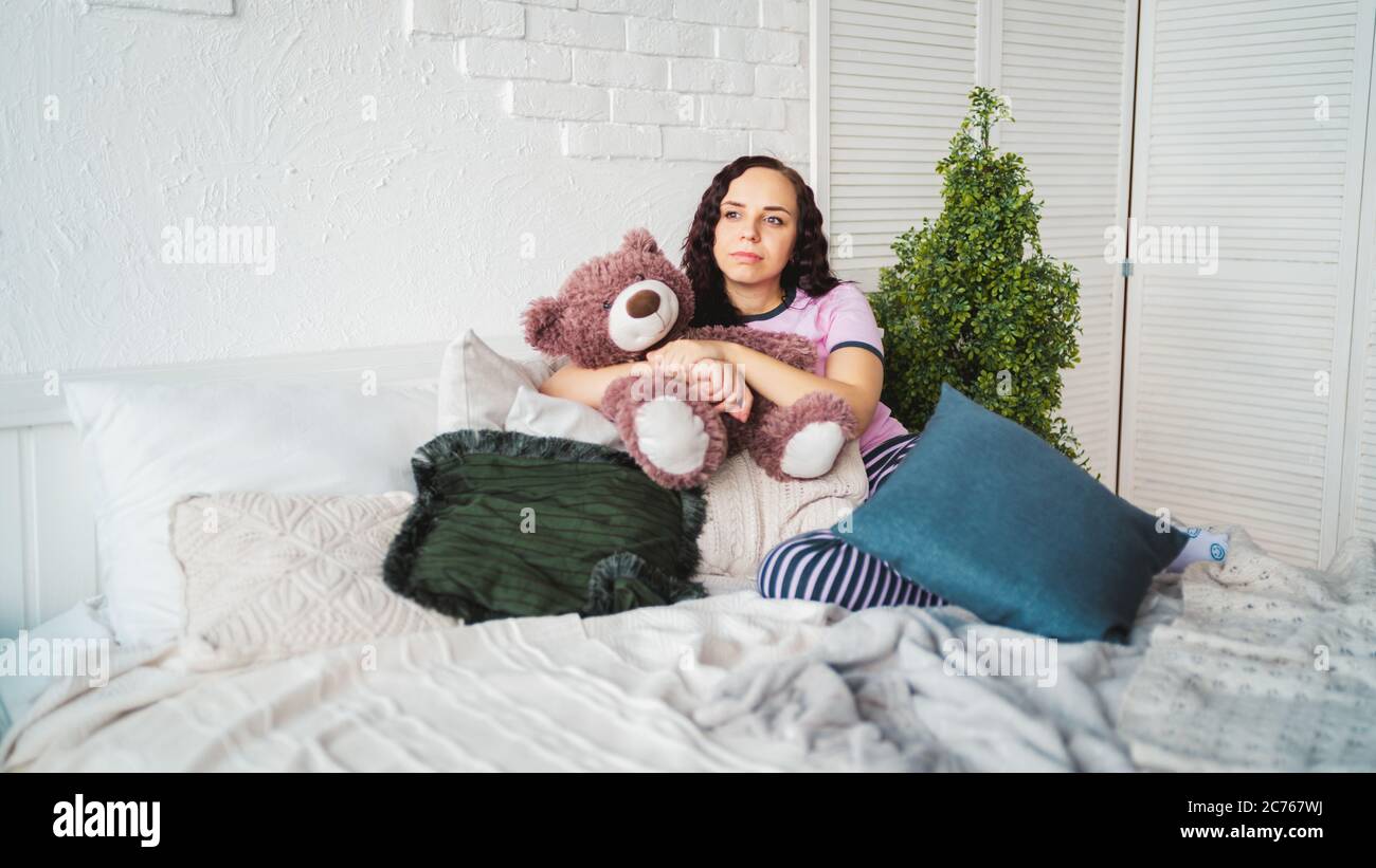 Junge Frau in Umarmung mit Spielzeug im Schlafzimmer ruhen. Erwachsene Frau im Schlafanzug liegend nachdenklich auf Kissen im Bett. Stockfoto