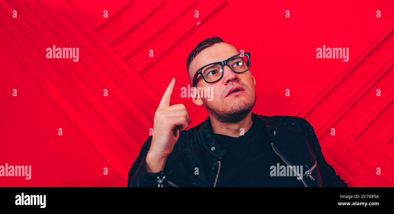 Hübscher junger Mann mit Brille hob seinen Zeigefinger nach oben von besucht durch Inspiration auf Hintergrund rote Wand. Das Konzept der großen Idee. Stockfoto
