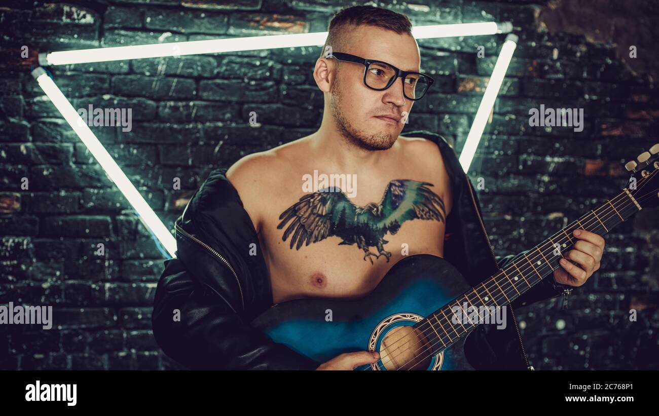 Tätowierter Mann spielt Gitarre in der Nähe von beleuchteten Wand. Stilvoller Kerl mit Vogel-Tattoo auf der Brust wegschauen und Gitarre spielen, während gegen schäbig stehen Stockfoto