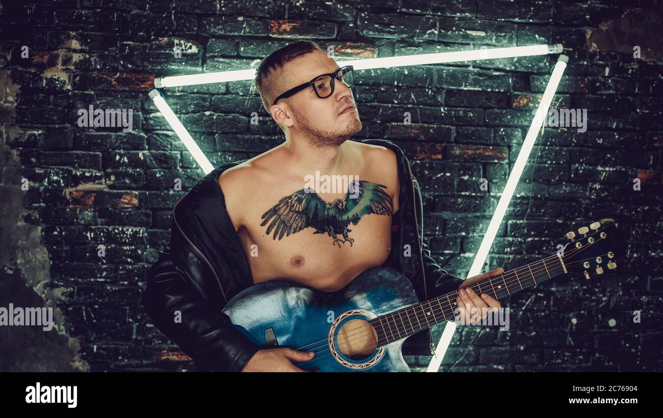 Tätowierter Mann spielt Gitarre in der Nähe von beleuchteten Wand. Stilvoller Kerl mit Vogel-Tattoo auf der Brust wegschauen und Gitarre spielen, während gegen schäbig stehen Stockfoto