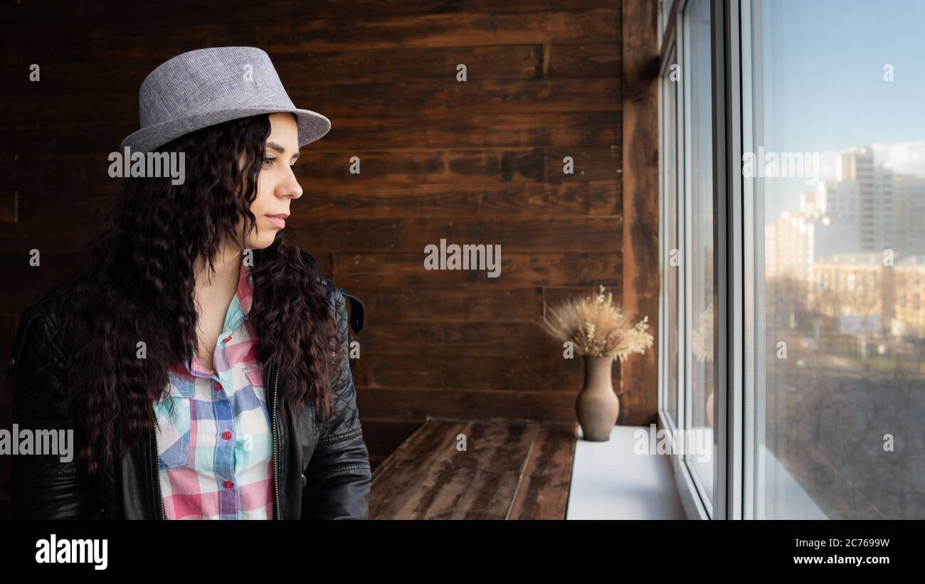 Junge schöne Frau in Hut und Lederjacke sitzen an den Fenstern. Porträt einer hübschen Frau, die tagsüber aus dem Fenster schaut. Stockfoto