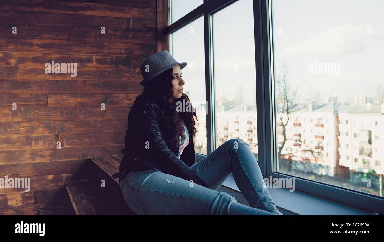 Junge schöne Frau in Hut und Lederjacke sitzen an den Fenstern. Porträt einer hübschen Frau, die tagsüber aus dem Fenster schaut. Stockfoto