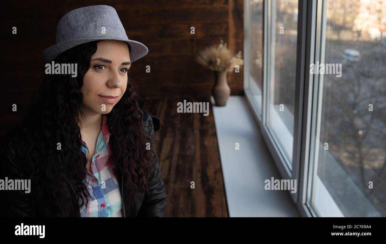Junge schöne Frau in Hut und Lederjacke sitzen an den Fenstern. Porträt einer hübschen Frau, die tagsüber aus dem Fenster schaut. Stockfoto