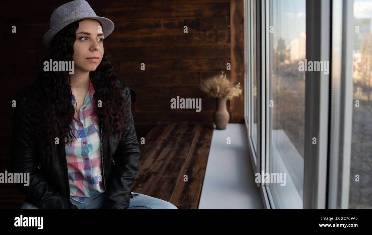 Junge schöne Frau in Hut und Lederjacke sitzen an den Fenstern. Porträt einer hübschen Frau, die tagsüber aus dem Fenster schaut. Stockfoto