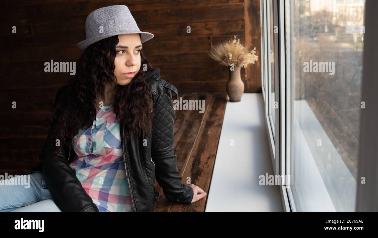 Junge schöne Frau in Hut und Lederjacke sitzen an den Fenstern. Porträt einer hübschen Frau, die tagsüber aus dem Fenster schaut. Stockfoto