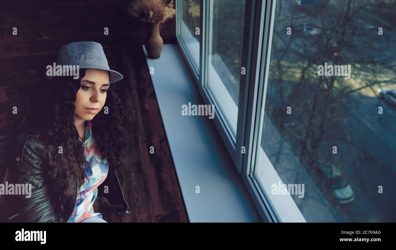 Junge schöne Frau in Hut und Lederjacke sitzen an den Fenstern. Porträt einer hübschen Frau, die tagsüber aus dem Fenster schaut. Stockfoto
