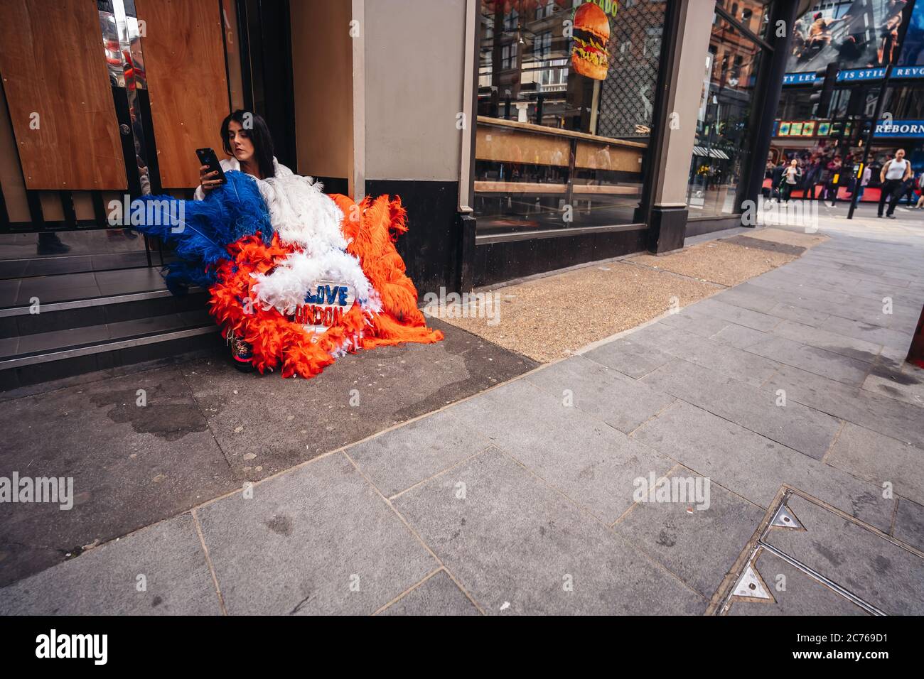 London / UK - 07/11/2020: Junge Dame mit 'London Love' Federfarbenes Kostüm sitzt auf dem Bürgersteig in SOHO Stockfoto