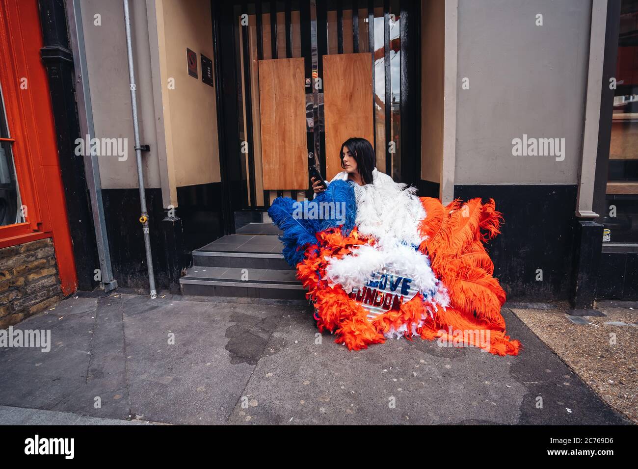 London / UK - 07/11/2020: Junge Dame mit 'London Love' Federfarbenes Kostüm sitzt auf dem Bürgersteig in SOHO Stockfoto