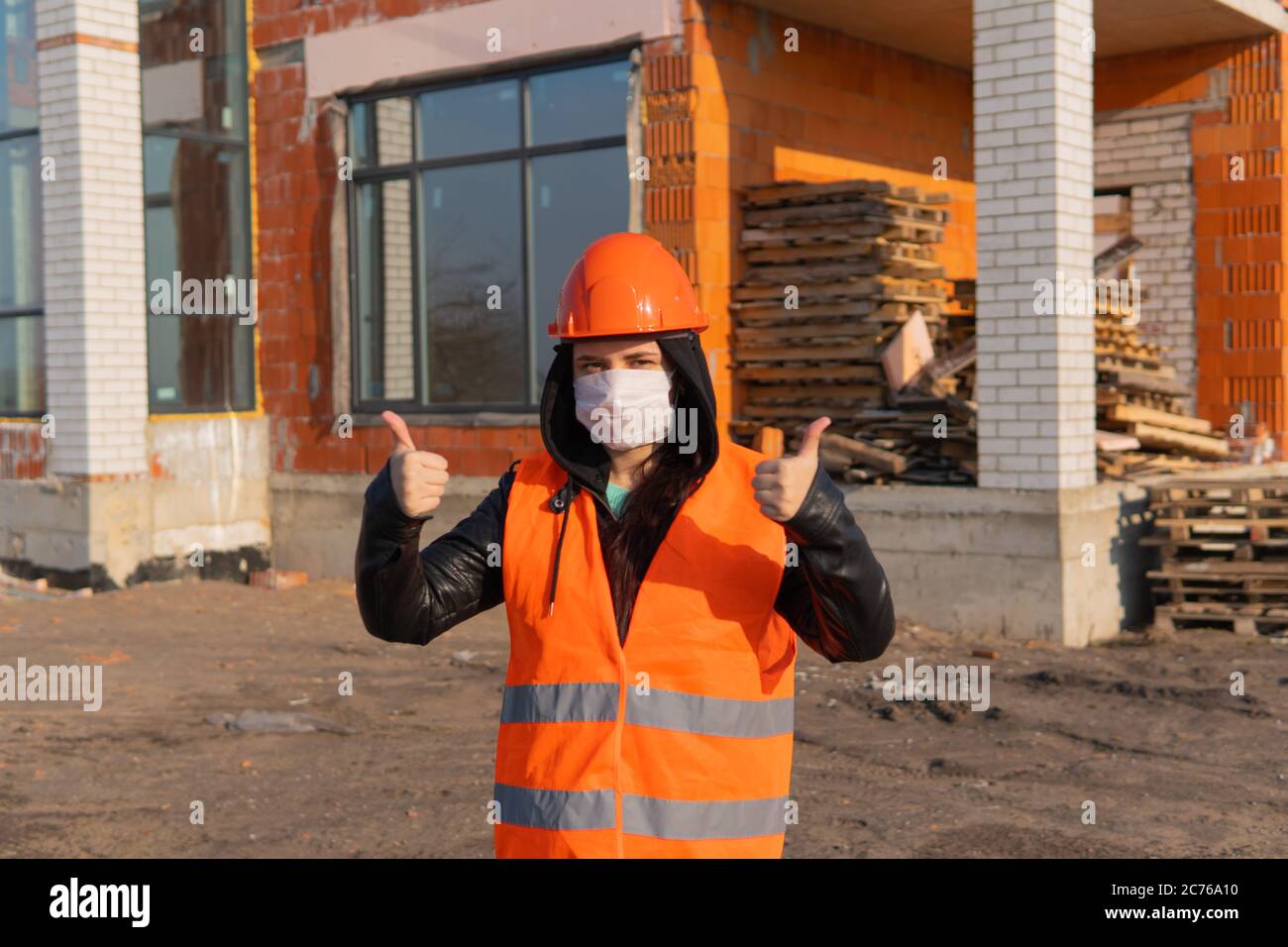 Bauarbeiterin in Overalls und in medizinischer Maske zeigt Daumen nach oben auf dem Hintergrund des Hauses im Bau. Junge Frau in Hut und Stockfoto