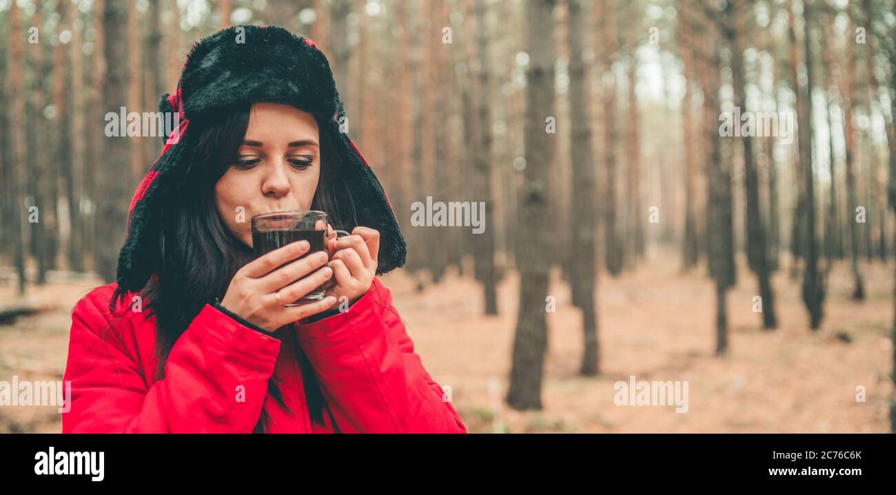 Ein Porträt der hübschen Frau mit einer Tasse Tee im Wald. Die Frau in einer Jacke und einem Hut mit Ohrenklappen trinkt bei kaltem Wetter einen Tee. Stockfoto