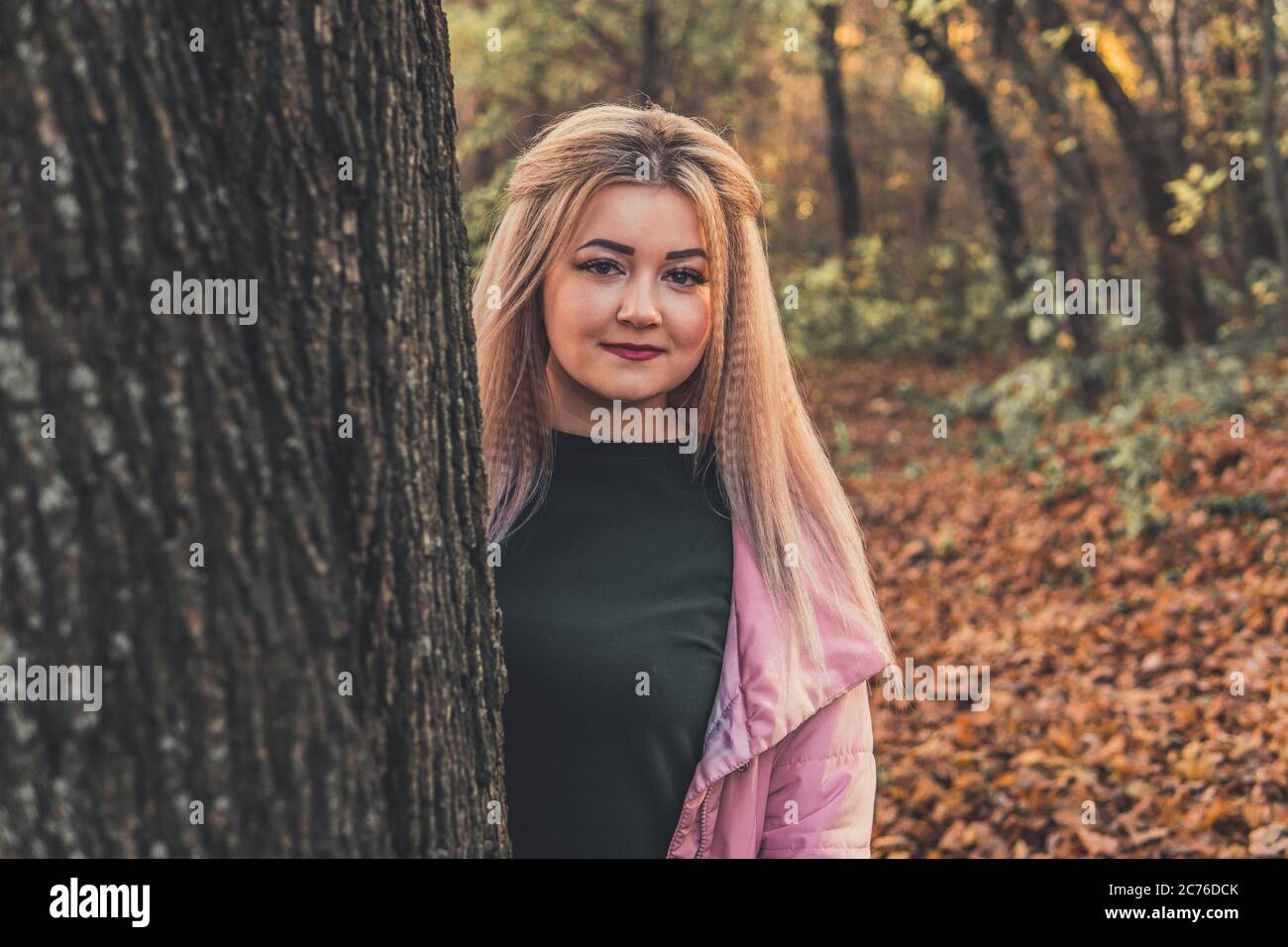 Schöne Frau, die hinter dem Baum herausschaut. Ziemlich ruhige Frau, die Baum berührt und die Kamera in einem ruhigen Wald anschaut Stockfoto