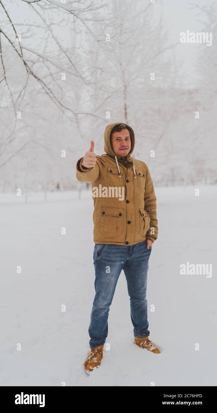 Schöner Mann, der im Winter im Park die Daumen hoch zeigt. Erwachsene junge Mann trägt beige Kapuzenjacke zeigt Geste der Zustimmung in Stockfoto