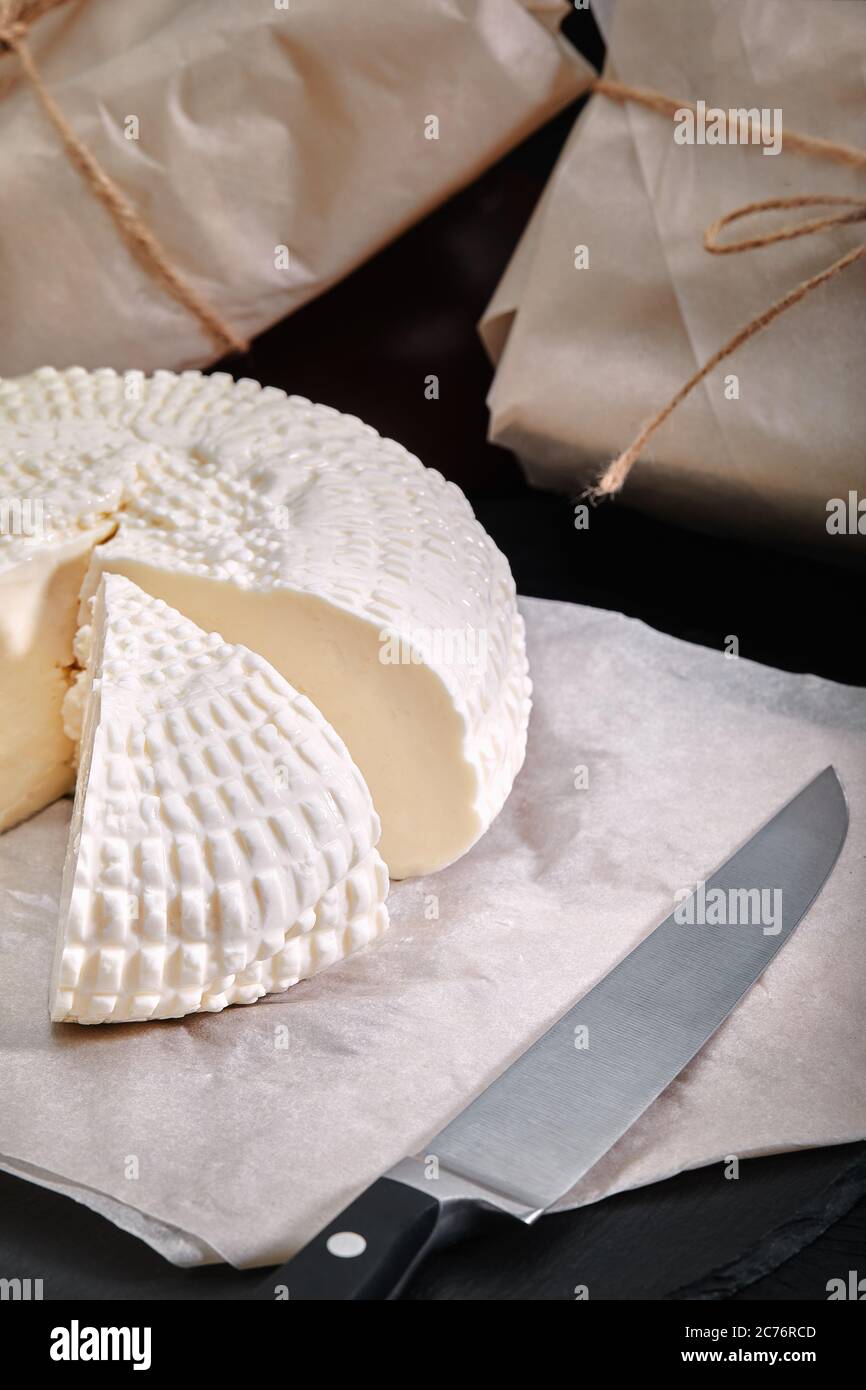 Adyghe Käse Schnitt ein dreieckiges Stück ab. In der Nähe befinden sich drei Käseköpfe, die in Papier verpackt sind. Auf einem Holzbrett. Hintergrund grauer Beton Stockfoto
