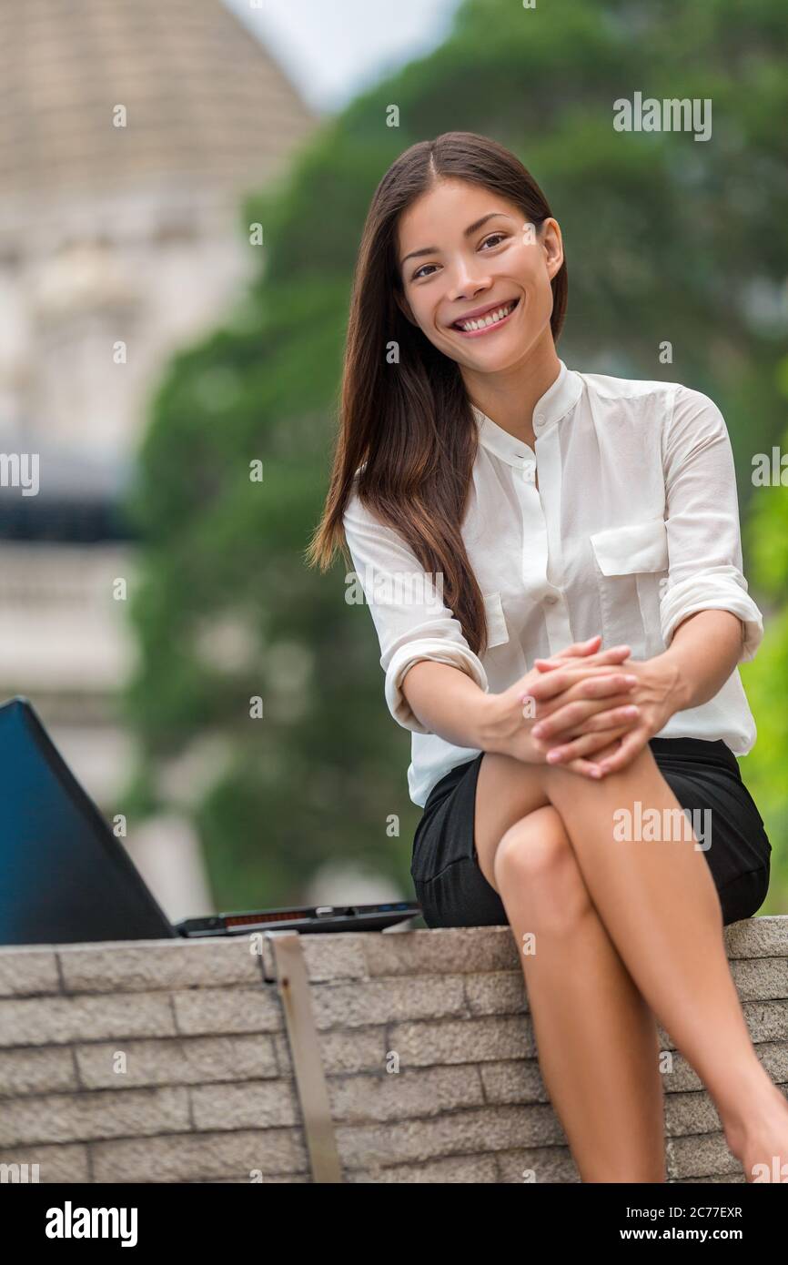 Glückliche multirassische asiatische junge Frau lächelt bei der Kamera auf der Arbeitspause im Stadtpark sitzen im Freien. Portrait von chinesischen kaukasischen Mädchen in ihren 20ern bei Stockfoto