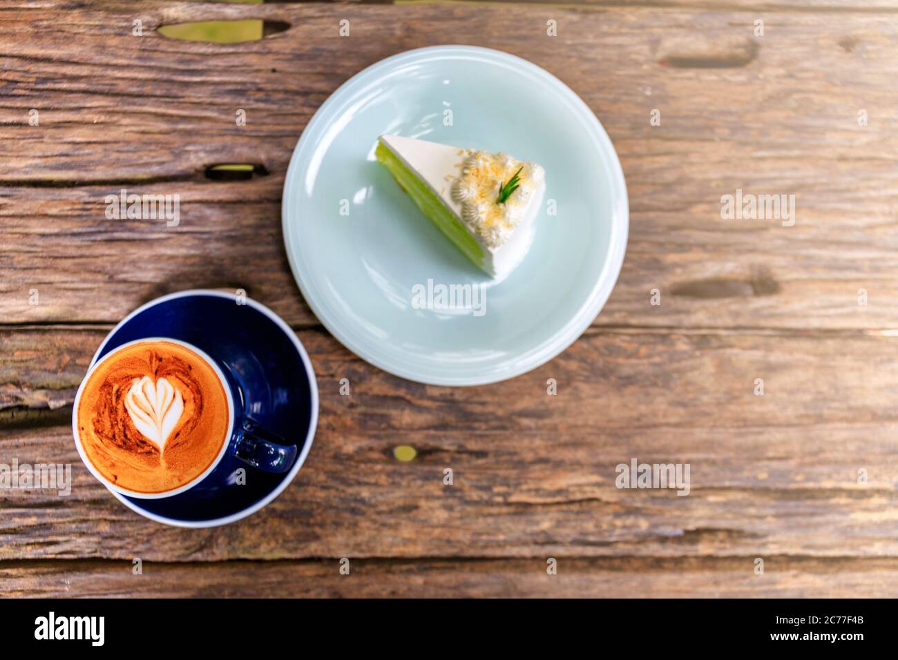 Draufsicht auf heißen Cappuccino-Kaffee und hausgemachten Kuchen im Café, herzförmiger Cappuccino-Kaffee in blauer Tasse auf einem Holztisch mit Kopierplatz. Stockfoto